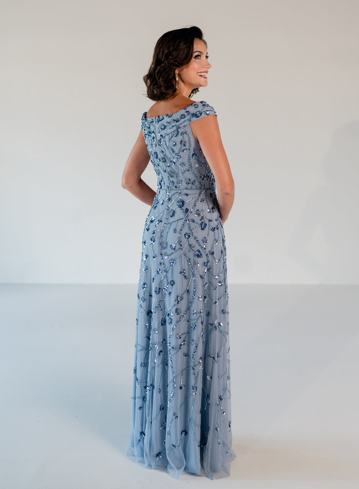 A woman modeling a floor-length light blue evening gown with intricate silver beaded floral patterns, photographed from the back against a plain white studio background. / Off the shoulder A-line gown with all over floral and trailing beading.