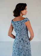 A woman shown from the back wearing a light blue off-the-shoulder evening gown with intricate blue and silver beadwork and sequins, set against a plain light gray studio background. / Off the shoulder A-line gown with all over floral and trailing beading.