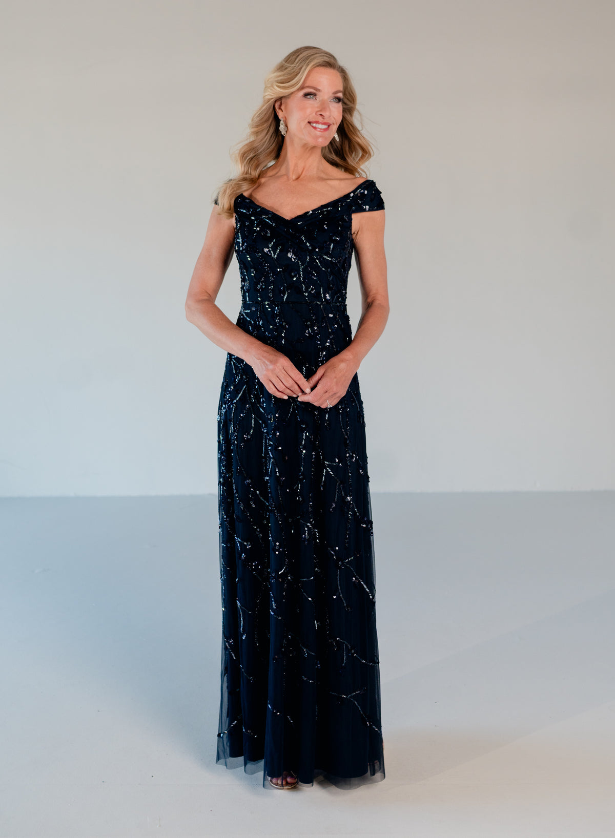 A woman modeling a floor-length off-the-shoulder gown in dark navy blue tulle with shimmering silver sequins, set against a plain light gray studio background. / Off the shoulder A-line gown with all over floral and trailing beading.