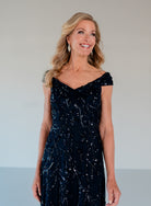 A smiling blonde woman wearing a dark navy off-the-shoulder evening gown with silver sequin details, standing against a neutral light gray background. / Off the shoulder A-line gown with all over floral and trailing beading.