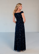 A woman modeling a floor-length off-the-shoulder evening gown in dark navy blue velvet with shimmering silver beadwork and tulle overlay, shown from the back against a plain light gray studio background. / Off the shoulder A-line gown with all over floral and trailing beading.