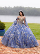 Woman in a blue and gold formal gown standing outdoors by a lake. / This ball gown features an embellished bodice and off-the-shoulder sleeves, paired with a full skirt decorated with intricate floral detailing and a large back bow.