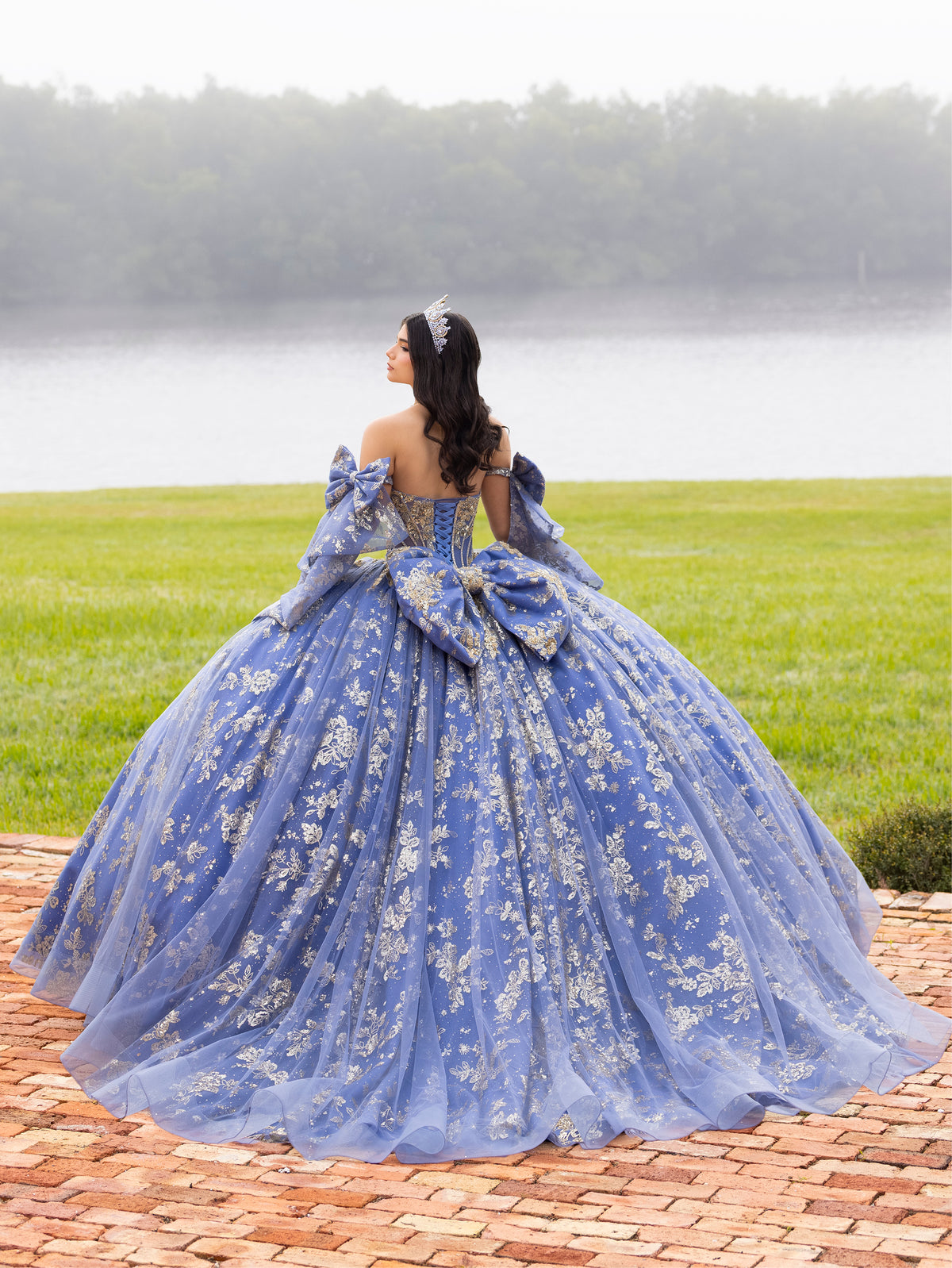 A woman wearing a voluminous blue ball gown with silver floral embroidery stands on a brick path outdoors, facing away from the camera toward a grassy field and body of water. / This ball gown features an embellished bodice and off-the-shoulder sleeves, paired with a full skirt decorated with intricate floral detailing and a large back bow.
