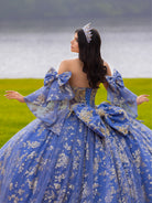 A woman wearing an elaborate royal blue ball gown with gold floral patterns, large bow details, and a matching tiara, photographed outdoors with a lake and green field in the background. / This ball gown features an embellished bodice and off-the-shoulder sleeves, paired with a full skirt decorated with intricate floral detailing and a large back bow.