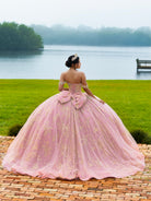 A woman stands outdoors by a lake wearing a voluminous light pink ball gown with gold floral embroidery, a large bow at the back, and off-the-shoulder straps. The background shows green grass, water, trees, and a brick path. / This ball gown features an embellished bodice and off-the-shoulder sleeves, paired with a full skirt decorated with intricate floral detailing and a large back bow.