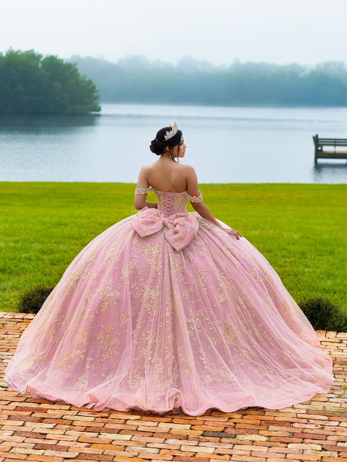 A woman stands outdoors by a lake wearing a voluminous light pink ball gown with gold floral embroidery, a large bow at the back, and off-the-shoulder straps. The background shows green grass, water, trees, and a brick path. / This ball gown features an embellished bodice and off-the-shoulder sleeves, paired with a full skirt decorated with intricate floral detailing and a large back bow.