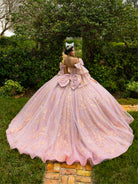 A woman stands outdoors in a lush garden wearing a voluminous light pink ball gown with gold floral embroidery, photographed from behind to show the back of the dress. The dress features a large bow at the waist and a full tulle skirt. / This ball gown features an embellished bodice and off-the-shoulder sleeves, paired with a full skirt decorated with intricate floral detailing and a large back bow.
