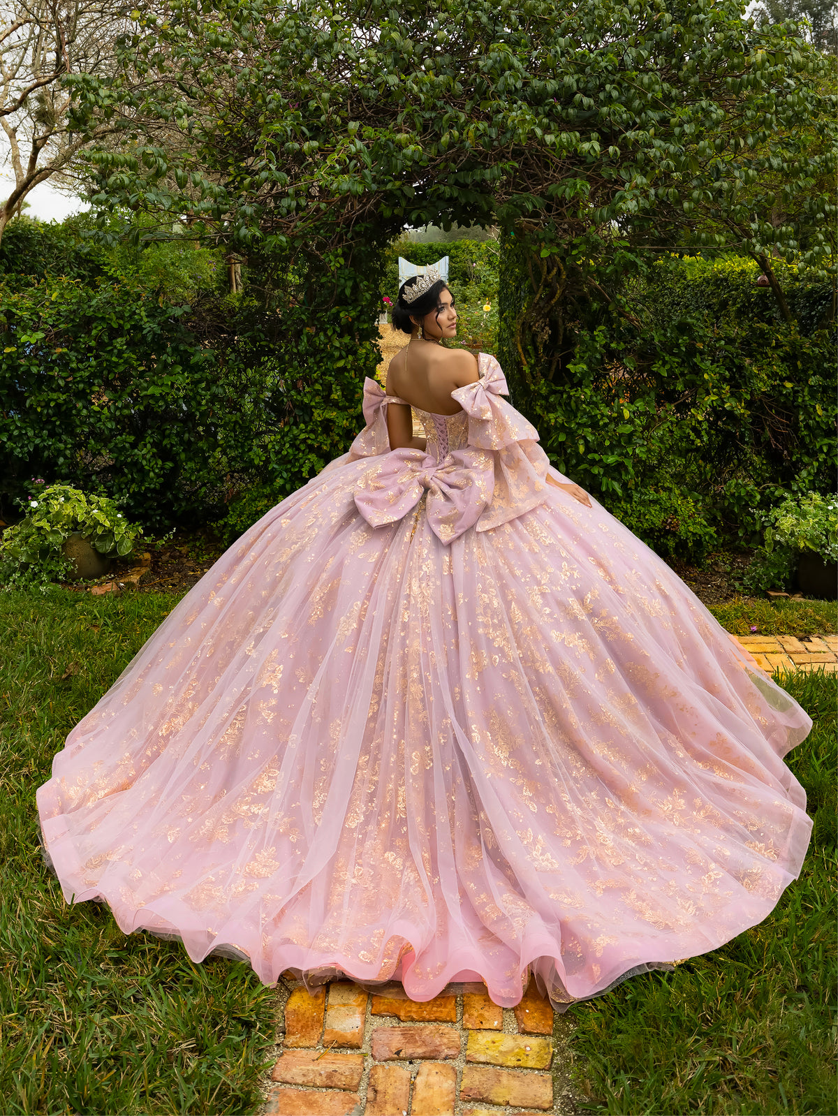 A woman stands outdoors in a lush garden wearing a voluminous light pink ball gown with gold floral embroidery, photographed from behind to show the back of the dress. The dress features a large bow at the waist and a full tulle skirt. / This ball gown features an embellished bodice and off-the-shoulder sleeves, paired with a full skirt decorated with intricate floral detailing and a large back bow.