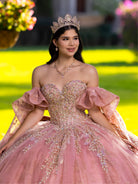 Woman in a pink, embellished dress with a tiara against a blurred outdoor background / This ball gown features an embellished bodice, off-the-shoulder sleeves, and a full skirt with floral detailing. A large bow at the back completes the look.