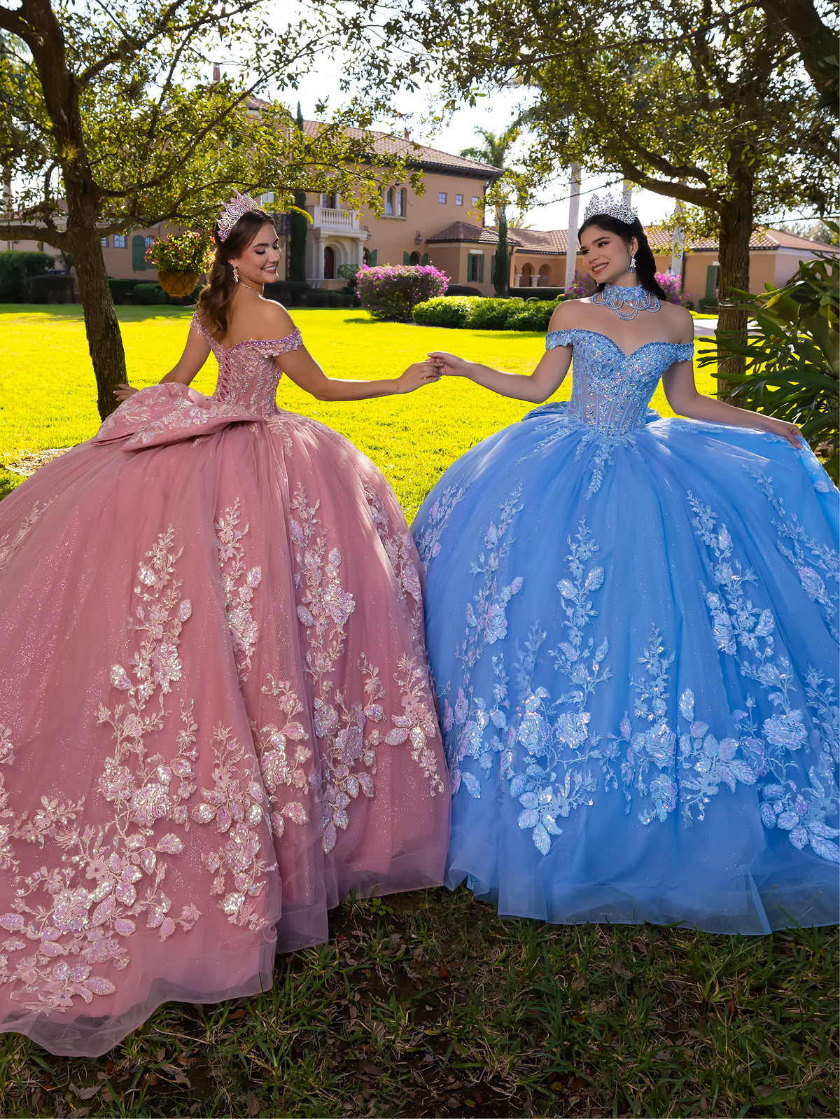 Two young women in off-the-shoulder ball gowns holding hands outdoors on a grassy lawn. One dress is dusty pink with white floral embroidery, and the other is sky blue with white floral embroidery. / This ball gown features an embellished bodice, off-the-shoulder sleeves, and a full skirt covered in detailed floral appliqué. The back is finished with a large decorative bow.