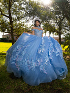 A woman wearing a voluminous light blue quinceañera ball gown with floral lace appliqués, standing outdoors in a grassy park with trees and sunlight in the background. / This ball gown features an embellished bodice, off-the-shoulder sleeves, and a full skirt covered in detailed floral appliqué. The back is finished with a large decorative bow.