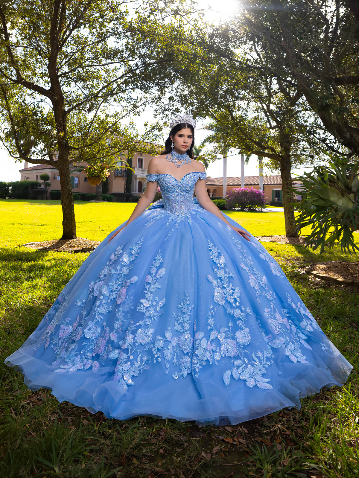 Woman in a blue floral gown standing outdoors with trees and a house in the background / This ball gown features an embellished bodice, off-the-shoulder sleeves, and a full skirt covered in detailed floral appliqué. The back is finished with a large decorative bow.