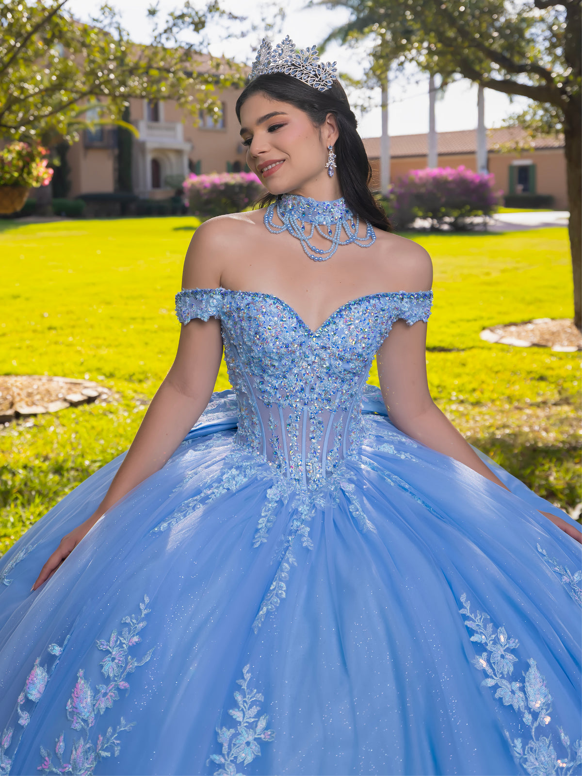 A young woman wearing a voluminous light blue quinceañera ball gown with floral embroidery, a matching beaded choker, and a silver tiara, posing outdoors on a green lawn with houses in the background. / This ball gown features an embellished bodice, off-the-shoulder sleeves, and a full skirt covered in detailed floral appliqué. The back is finished with a large decorative bow.