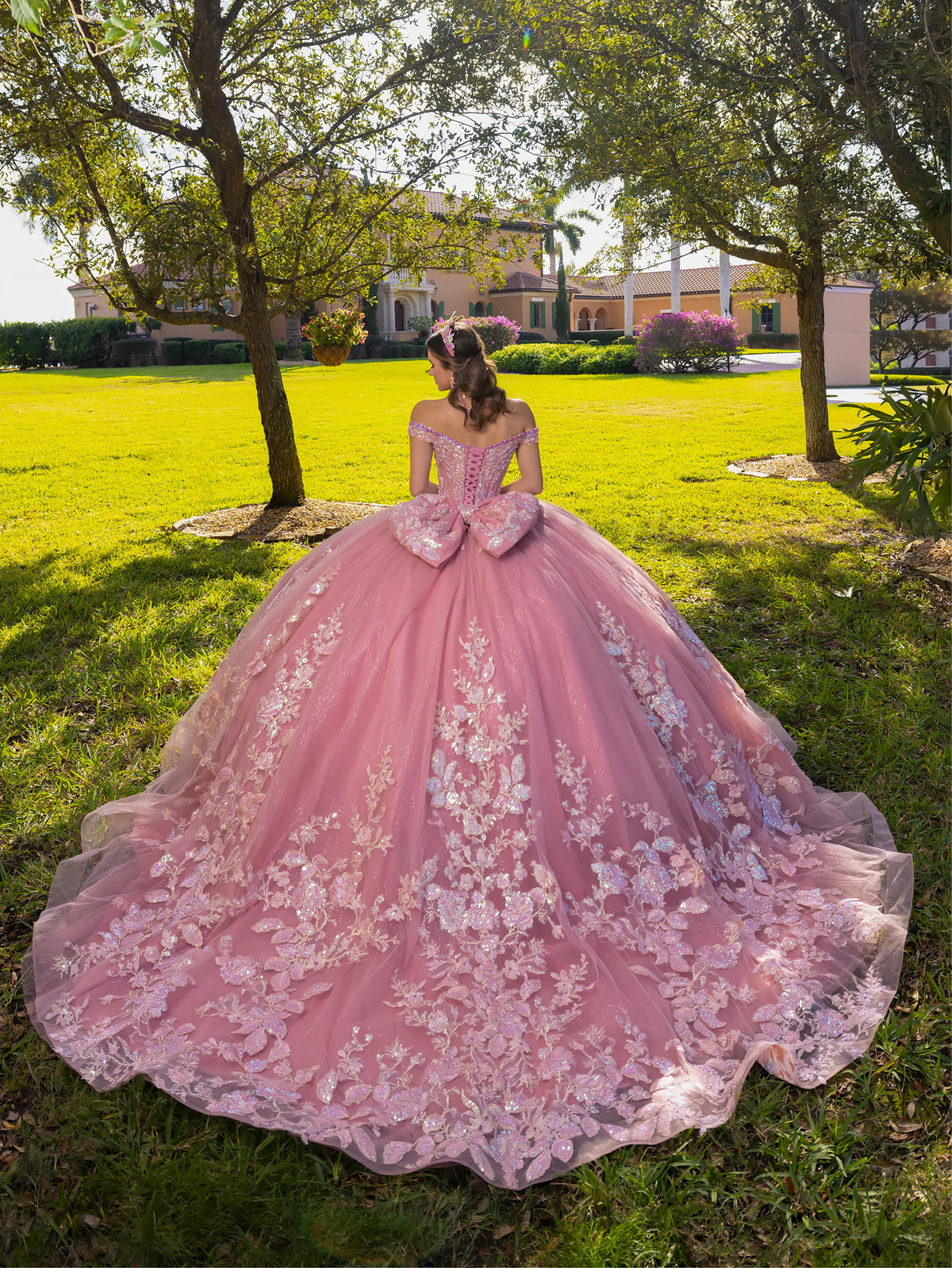 A woman stands outdoors on a grassy lawn wearing an elaborate pink ball gown with floral lace appliqué, a fitted off-the-shoulder bodice, and a very full skirt with a long train. / This ball gown features an embellished bodice, off-the-shoulder sleeves, and a full skirt covered in detailed floral appliqué. The back is finished with a large decorative bow.