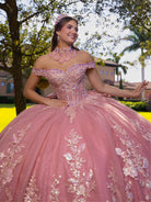 A woman wearing a voluminous pink ball gown with off-the-shoulder straps, a heavily embellished corset bodice, and large floral appliques on the skirt, posed outdoors in front of trees and houses. / This ball gown features an embellished bodice, off-the-shoulder sleeves, and a full skirt covered in detailed floral appliqué. The back is finished with a large decorative bow.