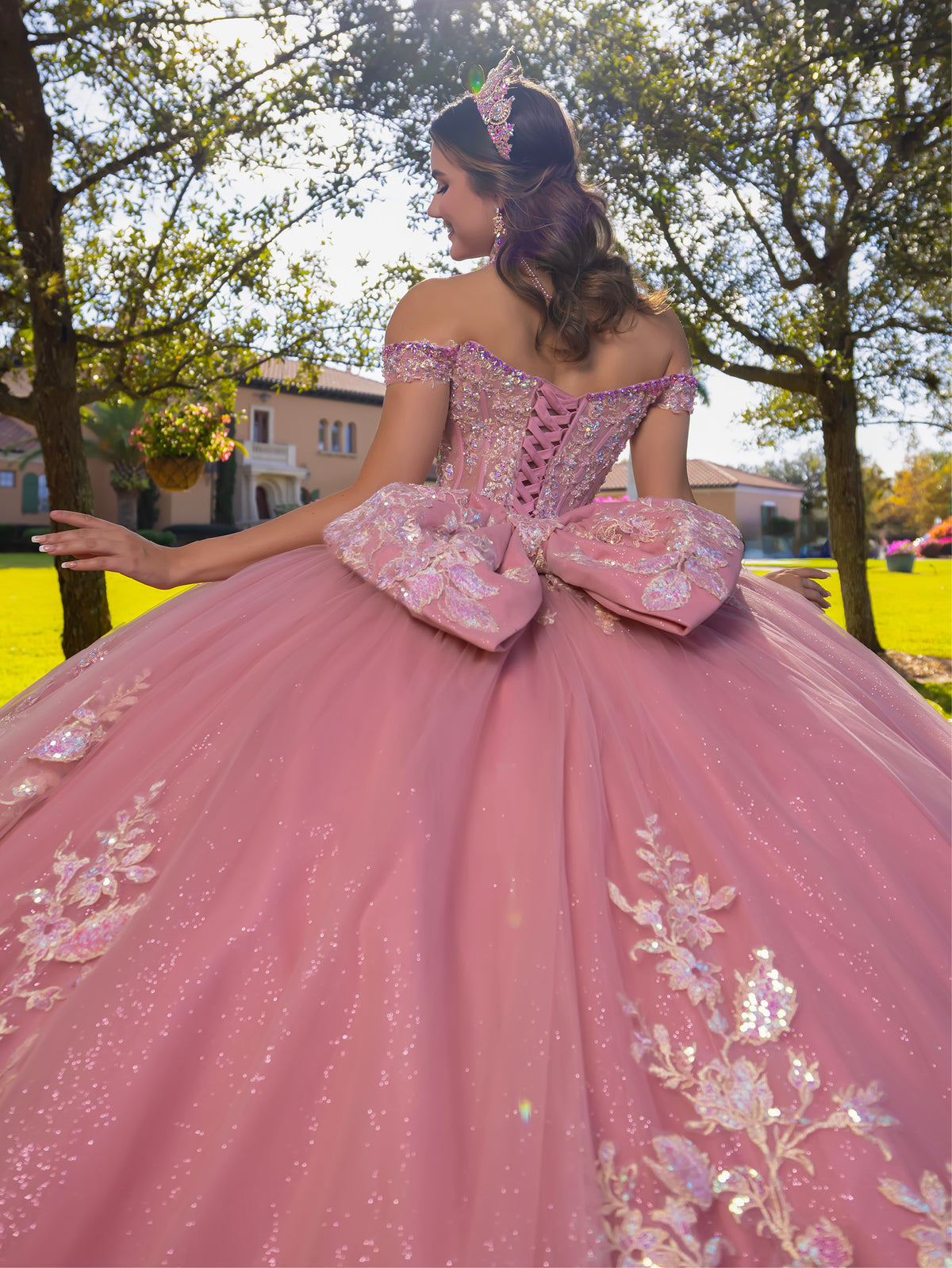 A woman wearing a voluminous pink ball gown with floral embroidery and large bow details, photographed outdoors among trees and grass. / This ball gown features an embellished bodice, off-the-shoulder sleeves, and a full skirt covered in detailed floral appliqué. The back is finished with a large decorative bow.