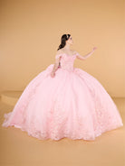 A woman modeling a voluminous light pink ball gown with off-the-shoulder straps, a fitted embellished bodice, and floral lace appliqué cascading down the tulle skirt, posed against a warm beige studio backdrop. / This ball gown features an embellished off-the-shoulder bodice and a full skirt with detailed floral accents.