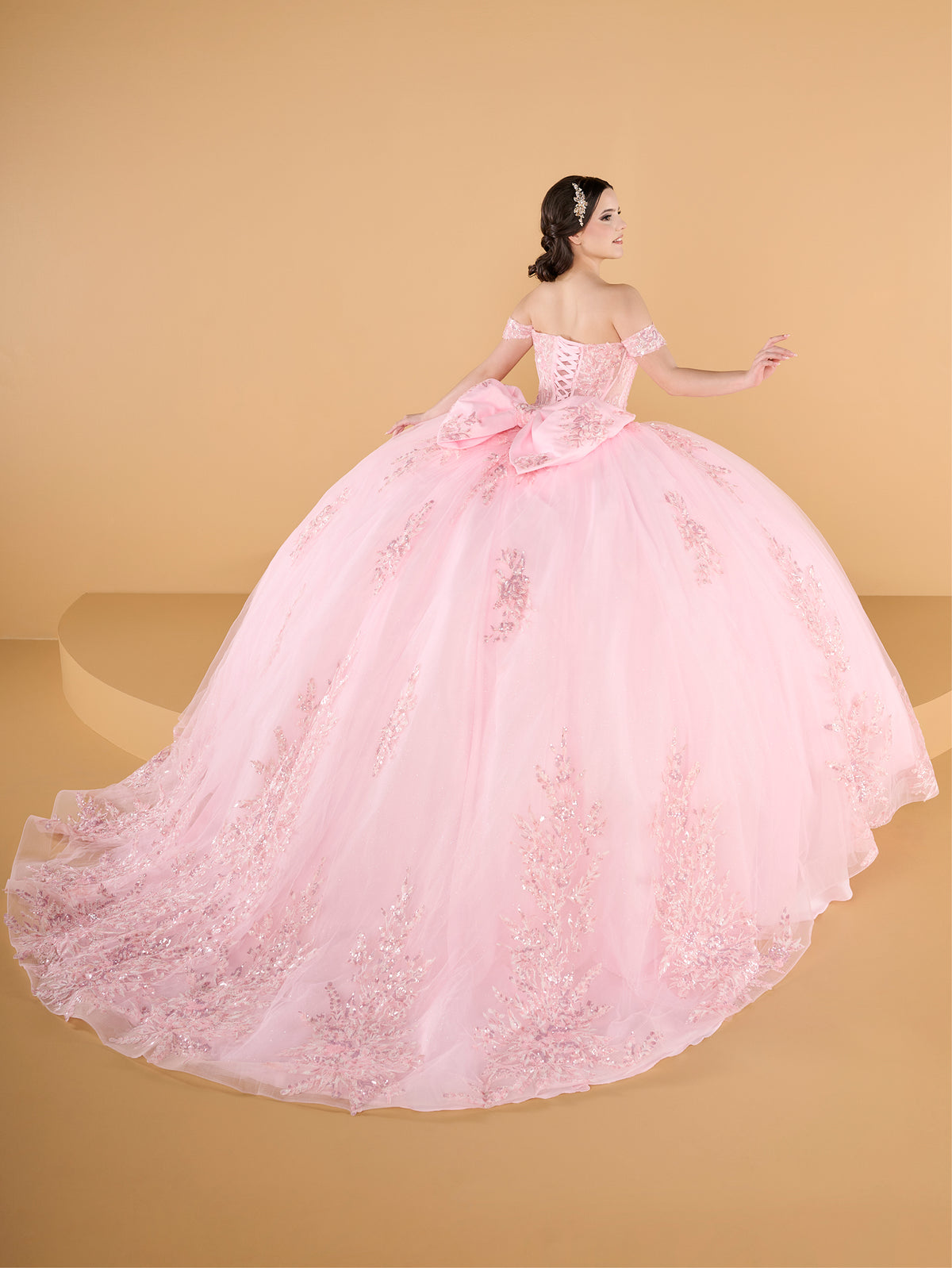 A woman modeling a voluminous light pink ball gown with floral lace appliques and a large bow at the waist, photographed against a warm beige studio backdrop. / This ball gown features an embellished off-the-shoulder bodice and a full skirt with detailed floral accents.