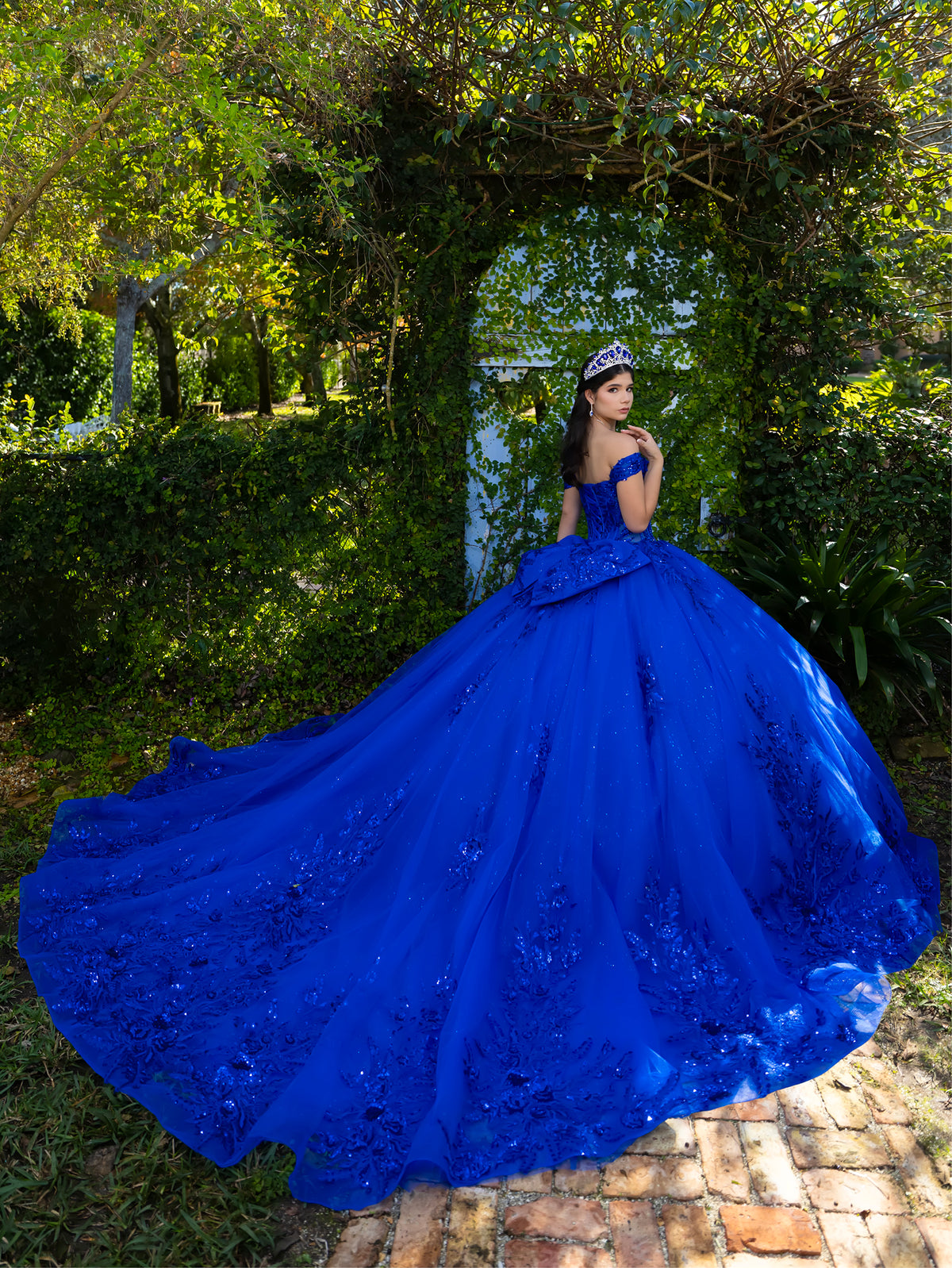 A woman wearing a voluminous royal blue ball gown with floral lace detailing stands outdoors in front of a leafy vine-covered archway on a brick path. / This ball gown features an embellished off-the-shoulder bodice and a full skirt with detailed floral accents.