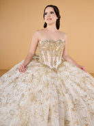 A young woman modeling a strapless ball gown with a heavily embellished gold beaded bodice and a voluminous cream tulle skirt decorated with gold floral embroidery and sequins, set against a solid warm beige background. / This ball gown features an embellished off-the-shoulder bodice, and a full skirt accented with detailed floral designs, finished with matching appliqué on the back.