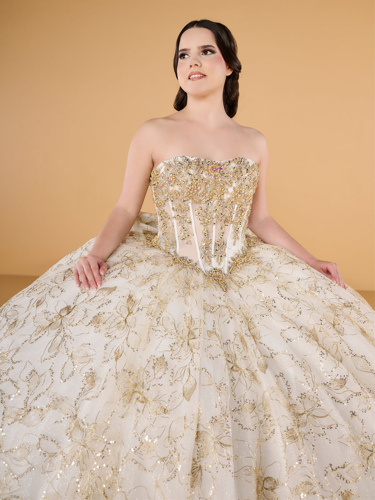 A young woman modeling a strapless ball gown with a heavily embellished gold beaded bodice and a voluminous cream tulle skirt decorated with gold floral embroidery and sequins, set against a solid warm beige background. / This ball gown features an embellished off-the-shoulder bodice, and a full skirt accented with detailed floral designs, finished with matching appliqué on the back.