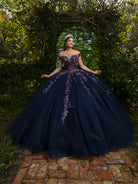 A woman wearing a voluminous off-the-shoulder navy ball gown with purple floral embroidery, standing outdoors on a brick path under a leafy archway. / This ball gown features an embellished bodice with off-the-shoulder sleeves and a full tulle skirt adorned with delicate floral appliqué. A large bow accents the back.