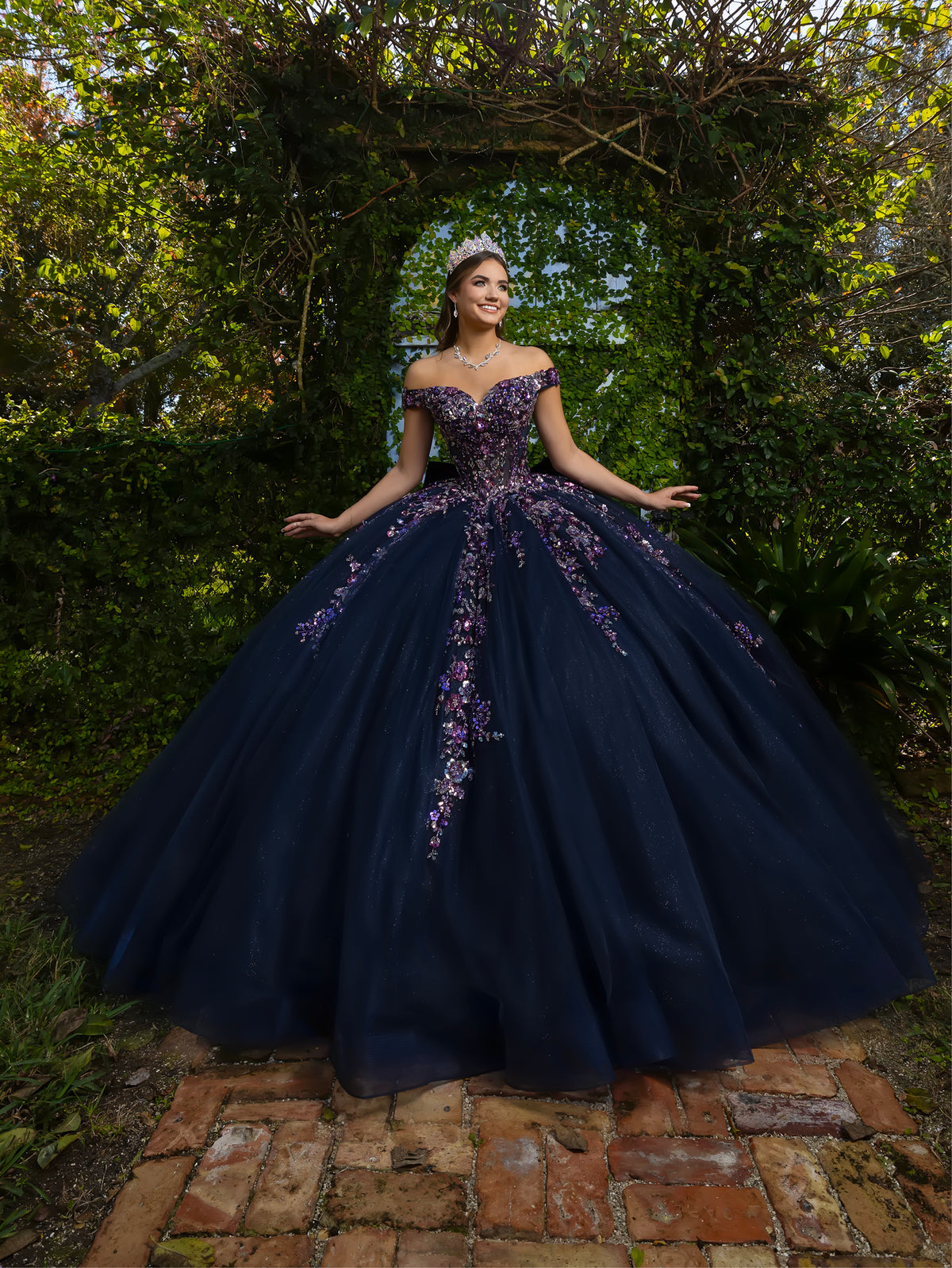 A woman wearing a voluminous off-the-shoulder navy ball gown with purple floral embroidery, standing outdoors on a brick path under a leafy archway. / This ball gown features an embellished bodice with off-the-shoulder sleeves and a full tulle skirt adorned with delicate floral appliqué. A large bow accents the back.