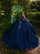 A woman stands outdoors in a lush garden wearing a voluminous off-the-shoulder ball gown. The dress is deep navy blue with purple floral embroidery and a large bow at the back, creating a dramatic train on the ground. / This ball gown features an embellished bodice with off-the-shoulder sleeves and a full tulle skirt adorned with delicate floral appliqué. A large bow accents the back.