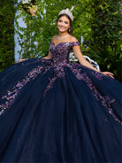 A woman wearing a voluminous off-the-shoulder ball gown in dark navy blue with purple and pink floral embroidery, seated outdoors against a green ivy-covered wall. / This ball gown features an embellished bodice with off-the-shoulder sleeves and a full tulle skirt adorned with delicate floral appliqué. A large bow accents the back.