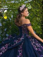 A woman wearing a dark navy off-the-shoulder ball gown with purple and pink floral sequin embellishments, posing outdoors against a green ivy-covered wall while wearing a jeweled tiara. / This ball gown features an embellished bodice with off-the-shoulder sleeves and a full tulle skirt adorned with delicate floral appliqué. A large bow accents the back.
