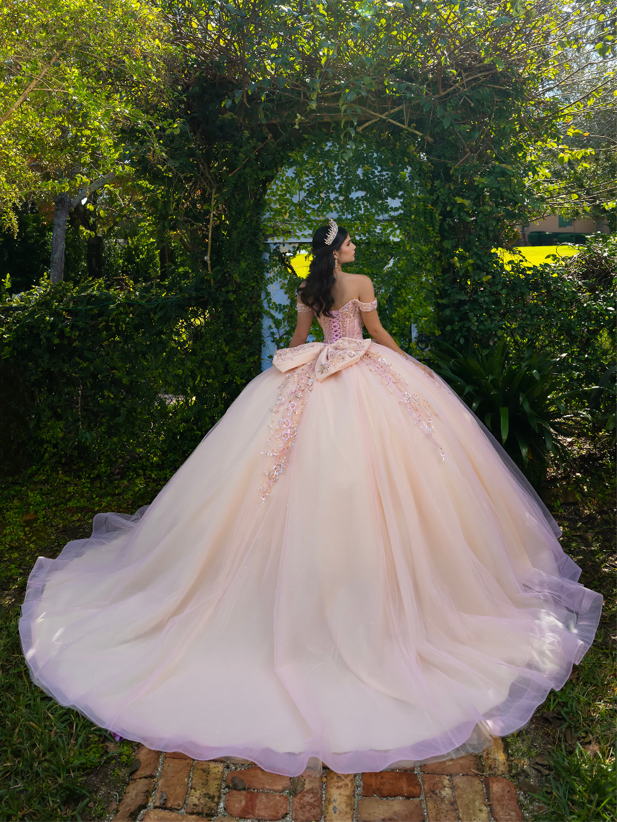 Woman in a pink gown standing in a garden with greenery around / This ball gown features an embellished bodice with off-the-shoulder sleeves and a full tulle skirt adorned with delicate floral appliqué. A large bow accents the back.