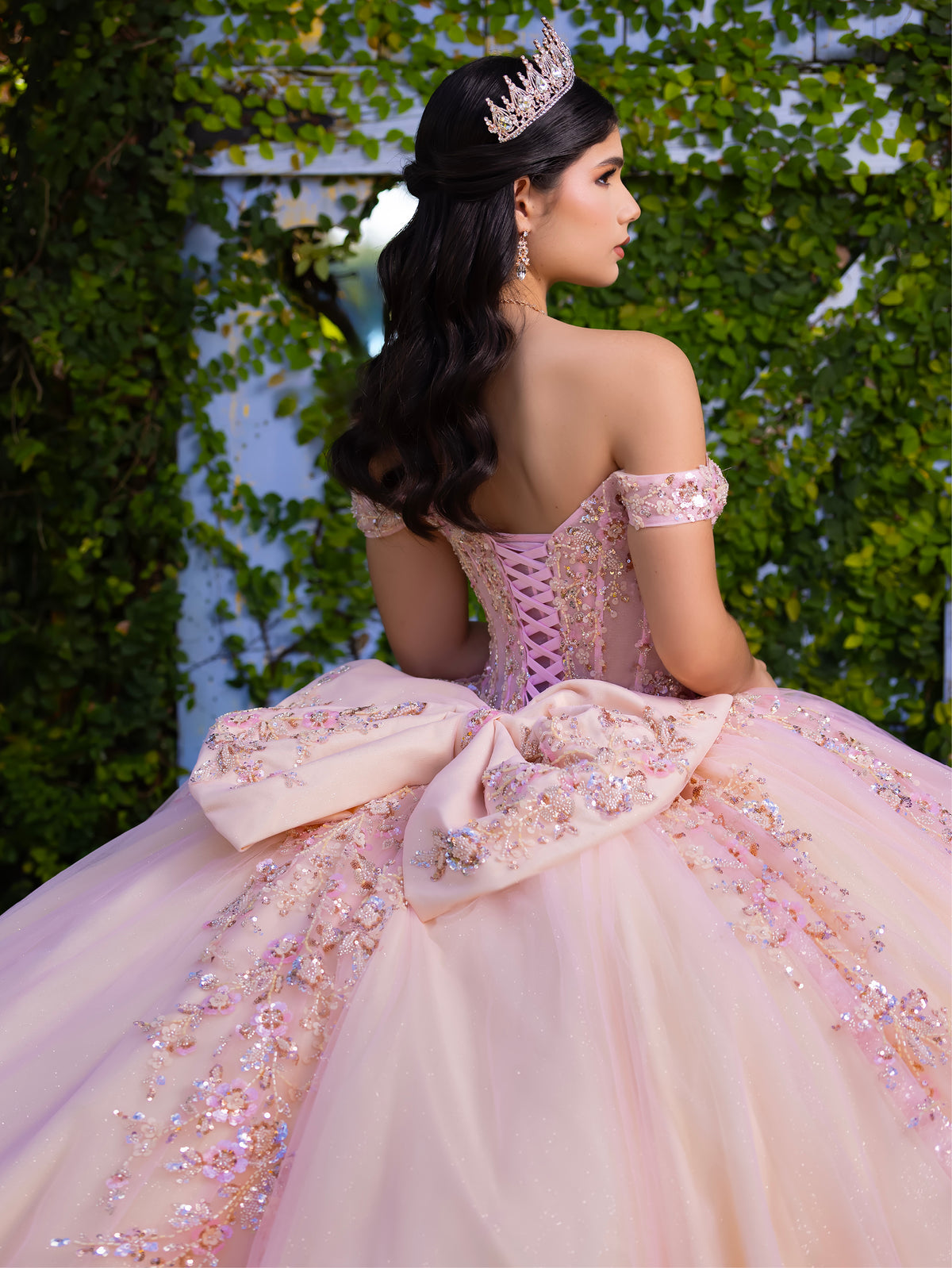 Woman in a pink quinceañera dress with intricate details, standing against a green leafy background. / This ball gown features an embellished bodice with off-the-shoulder sleeves and a full tulle skirt adorned with delicate floral appliqué. A large bow accents the back.