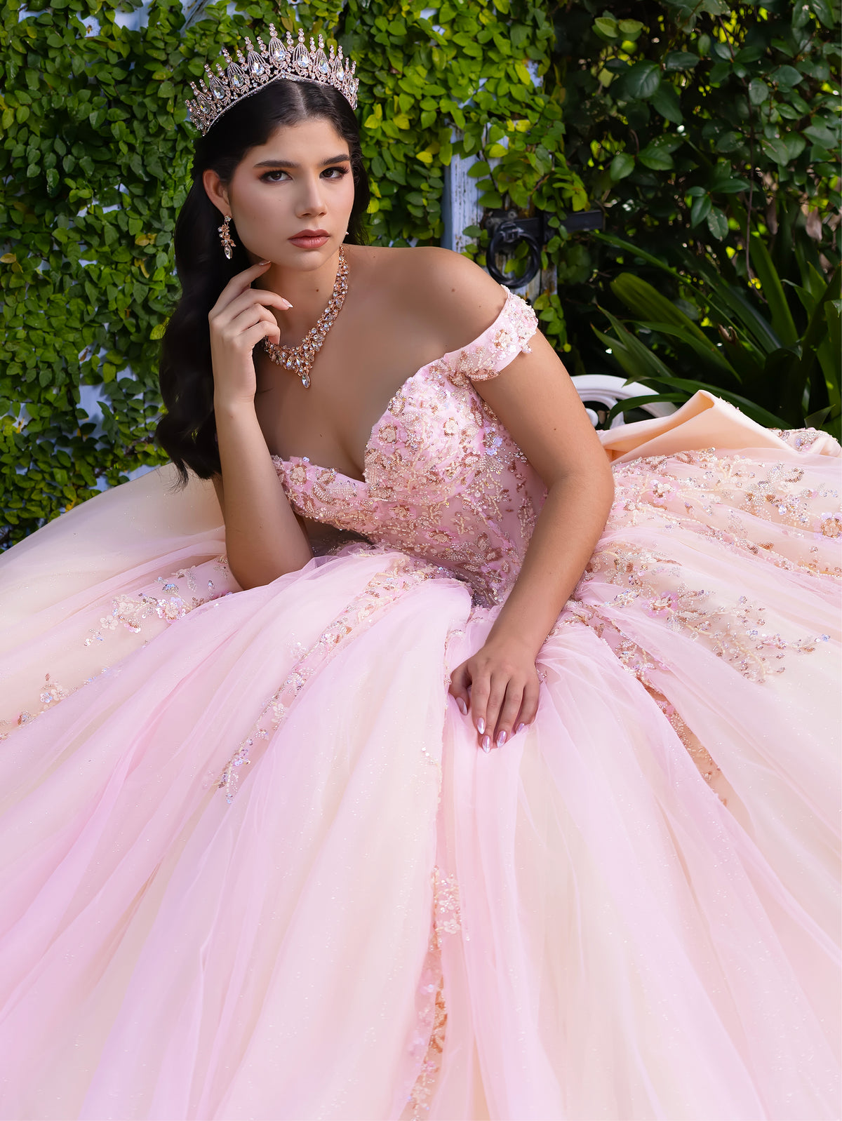 Woman in a pink gown with floral details and a tiara, standing outdoors. / This ball gown features an embellished bodice with off-the-shoulder sleeves and a full tulle skirt adorned with delicate floral appliqué. A large bow accents the back.