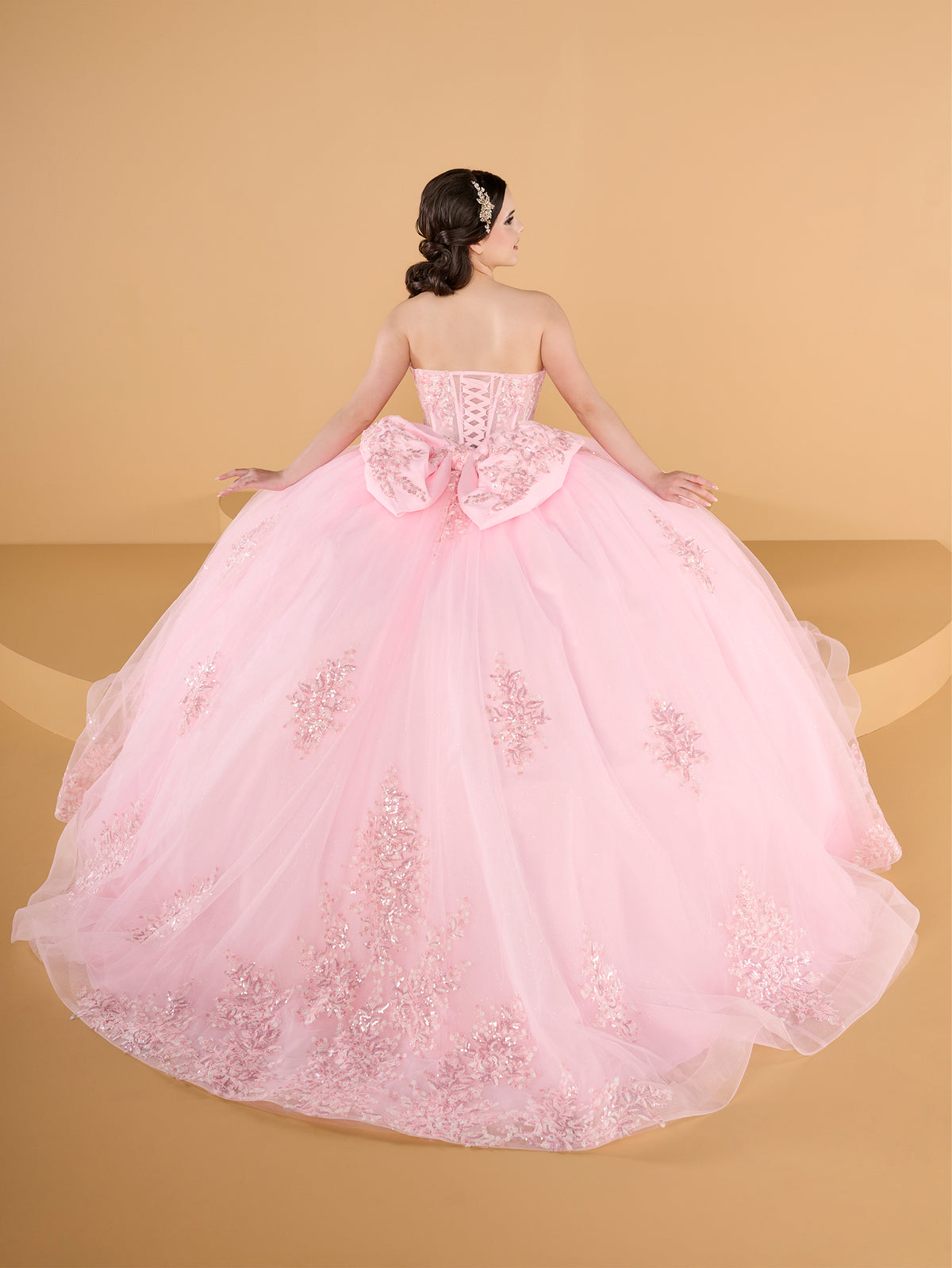 A pink strapless ball gown with a sweetheart neckline, lace-up corset-style bodice, and a large bow at the waist, shown from the back on a model against a warm beige studio backdrop. / This ball gown features a fitted, embellished bodice and a full skirt decorated with floral appliqué, finished with a bow at the back.