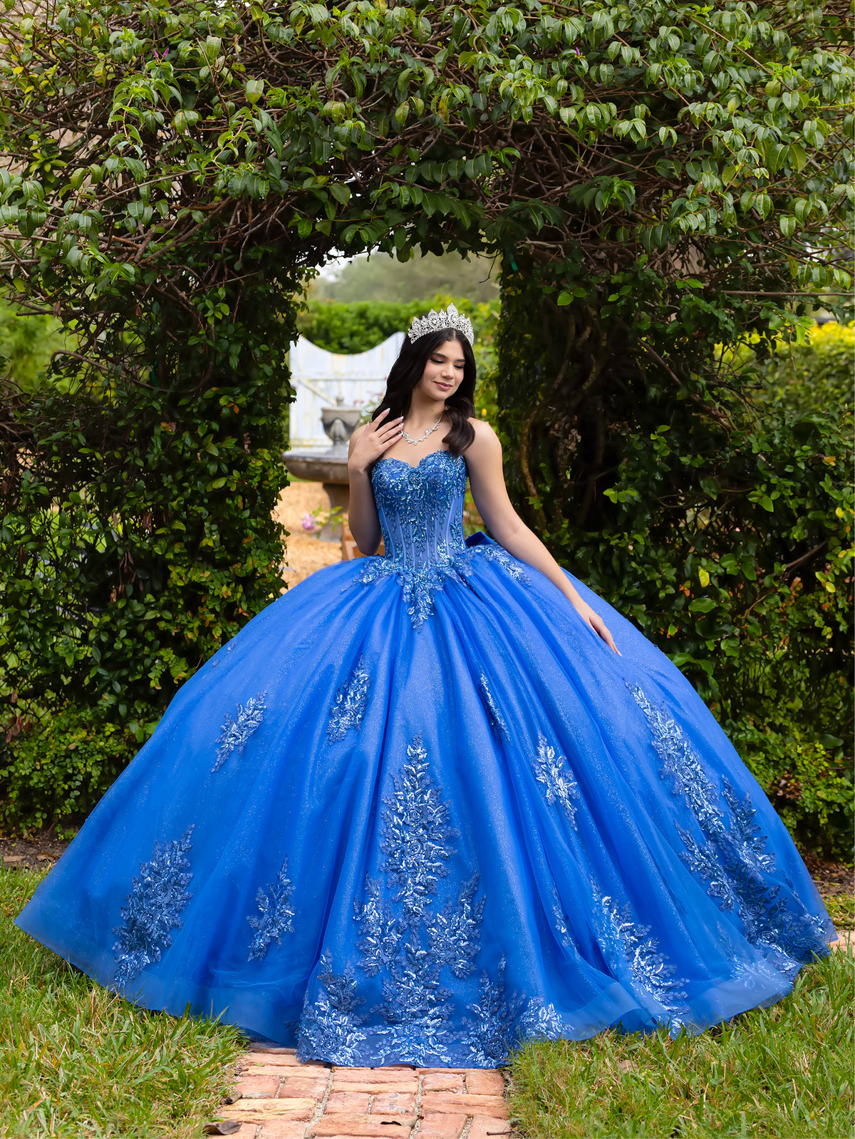 A woman wearing a voluminous royal blue ball gown with silver floral embroidery stands outdoors on a brick path framed by green hedges and trees. / This ball gown features a fitted, embellished bodice and a full skirt decorated with floral appliqué, finished with a bow at the back.