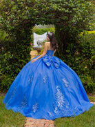 A young woman wearing a voluminous royal blue ball gown with silver floral embroidery and a large bow at the back, photographed outdoors in a garden setting. / This ball gown features a fitted, embellished bodice and a full skirt decorated with floral appliqué, finished with a bow at the back.