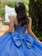 A young woman seen from behind wearing a royal blue strapless ball gown with a large bow at the back, lace-up corset bodice, and silver tiara in an outdoor garden setting. / This ball gown features a fitted, embellished bodice and a full skirt decorated with floral appliqué, finished with a bow at the back.