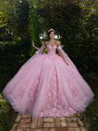 A woman wearing a voluminous light pink ball gown with floral appliqué and off-the-shoulder tulle sleeves, standing outdoors on a brick path surrounded by green foliage. / This ball gown features a floral-embellished bodice and a full tulle skirt with cascading 3D flowers, finished with a decorative bow at the back.