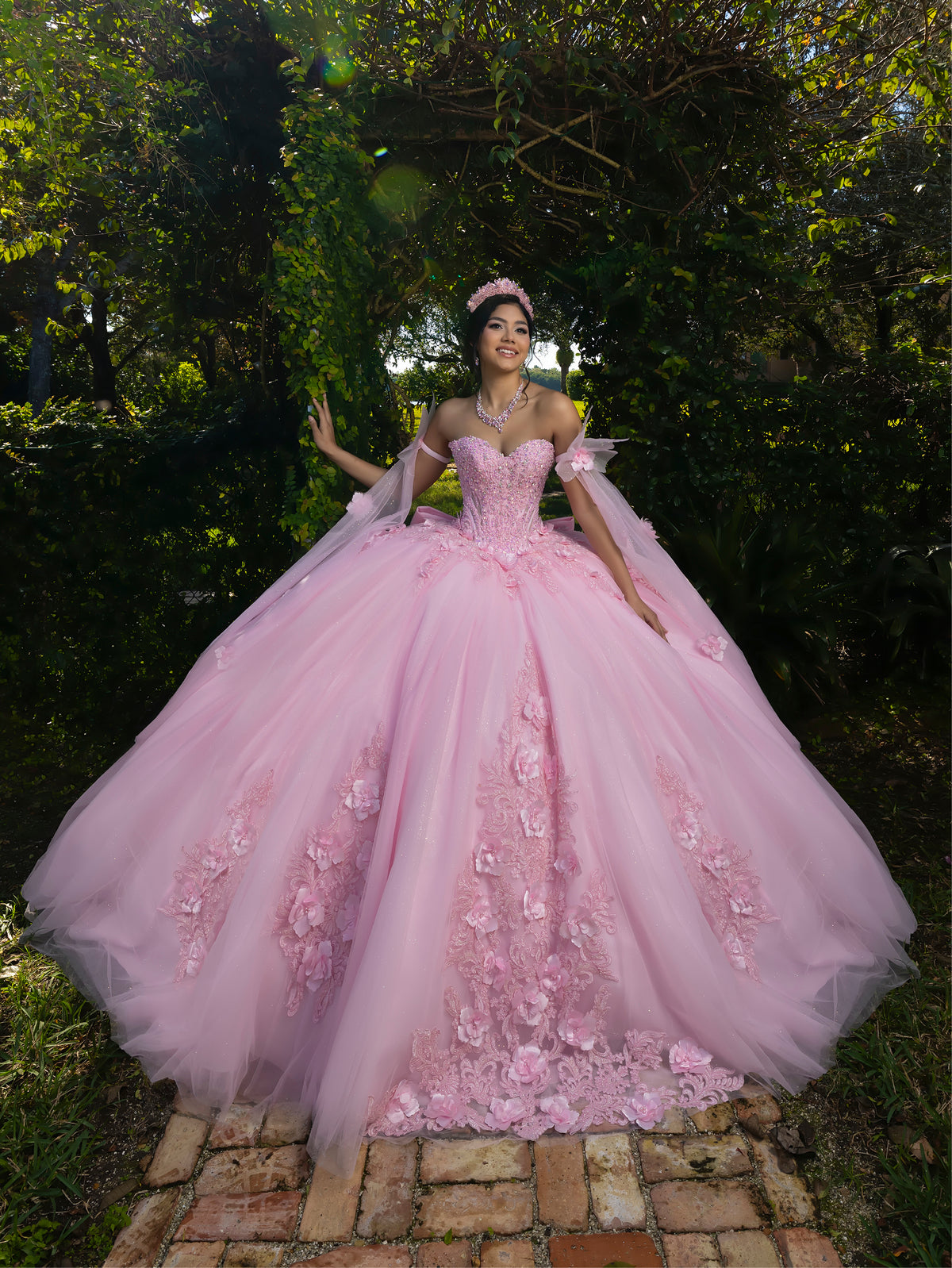 A woman wearing a voluminous light pink ball gown with floral appliqué and off-the-shoulder tulle sleeves, standing outdoors on a brick path surrounded by green foliage. / This ball gown features a floral-embellished bodice and a full tulle skirt with cascading 3D flowers, finished with a decorative bow at the back.