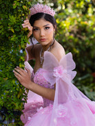 A young woman wearing a pink quinceañera dress with a tiara, posing outdoors next to a leafy green vine wall. / This ball gown features a floral-embellished bodice and a full tulle skirt with cascading 3D flowers, finished with a decorative bow at the back.