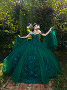 A woman stands outdoors in a lush garden wearing an elaborate emerald green ball gown with floral embroidery and sheer draped sleeves. The background features dense green foliage and a brick path. / This ball gown features a floral-embellished bodice and a full tulle skirt with cascading 3D flowers, finished with a decorative bow at the back.