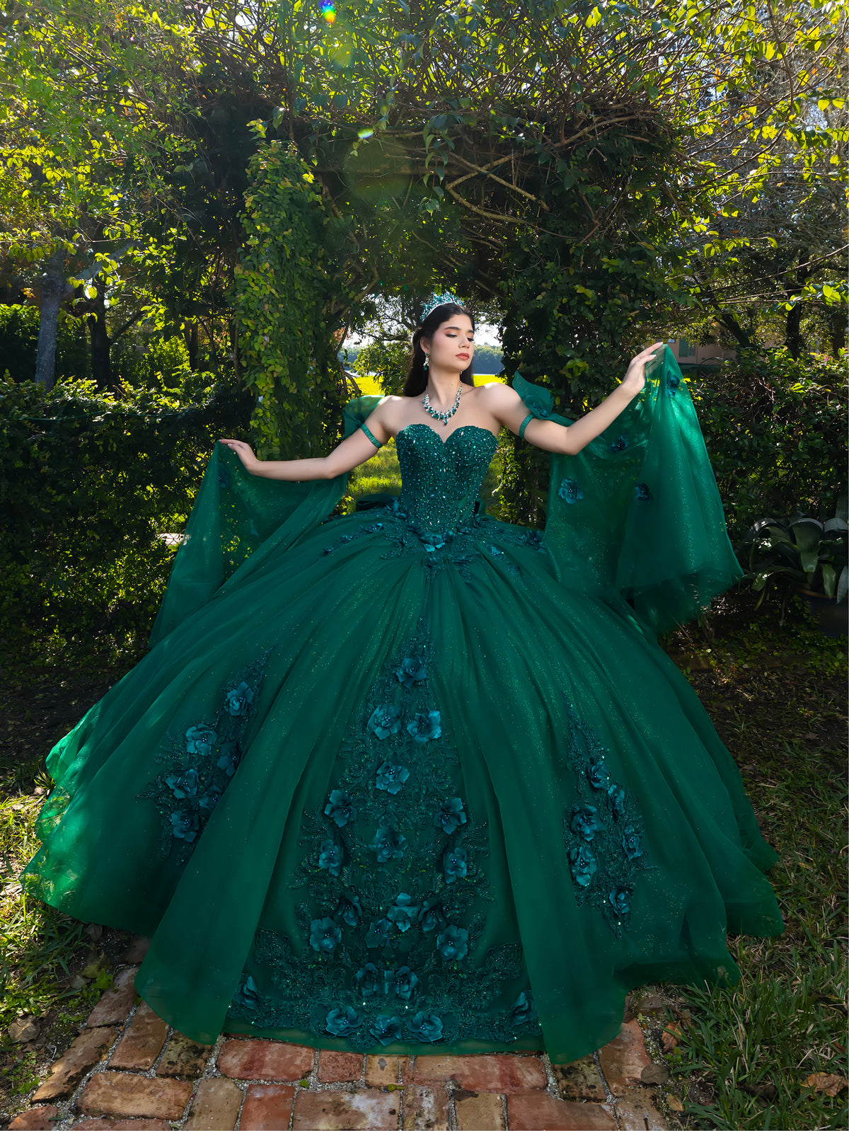A woman stands outdoors in a lush garden wearing an elaborate emerald green ball gown with floral embroidery and sheer draped sleeves. The background features dense green foliage and a brick path. / This ball gown features a floral-embellished bodice and a full tulle skirt with cascading 3D flowers, finished with a decorative bow at the back.