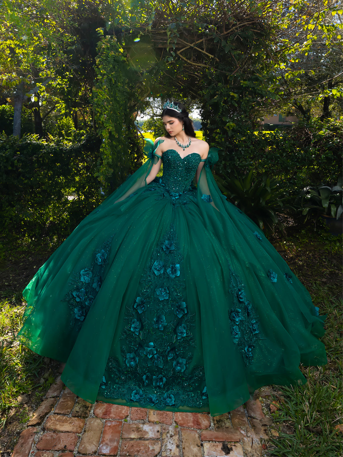 A woman stands outdoors in a lush garden wearing an elaborate emerald green ball gown with floral appliqué, sheer draped sleeves, and a voluminous tulle skirt. / This ball gown features a floral-embellished bodice and a full tulle skirt with cascading 3D flowers, finished with a decorative bow at the back.