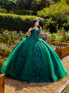 A woman wearing a voluminous emerald green ball gown with floral lace appliques, posed outdoors in a garden setting with gravel and greenery in the background. / This ball gown features a floral-embellished bodice and a full tulle skirt with cascading 3D flowers, finished with a decorative bow at the back.