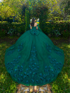 A woman stands outdoors in a lush garden wearing a dramatic emerald green ball gown from behind, showing an enormous train with 3D floral appliqués and a large bow at the back of the neckline. / This ball gown features a floral-embellished bodice and a full tulle skirt with cascading 3D flowers, finished with a decorative bow at the back.
