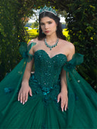A woman wearing an emerald green strapless ball gown with sequined embellishments, floral appliqué, and sheer draped fabric, posed outdoors in front of a leafy green archway. / This ball gown features a floral-embellished bodice and a full tulle skirt with cascading 3D flowers, finished with a decorative bow at the back.