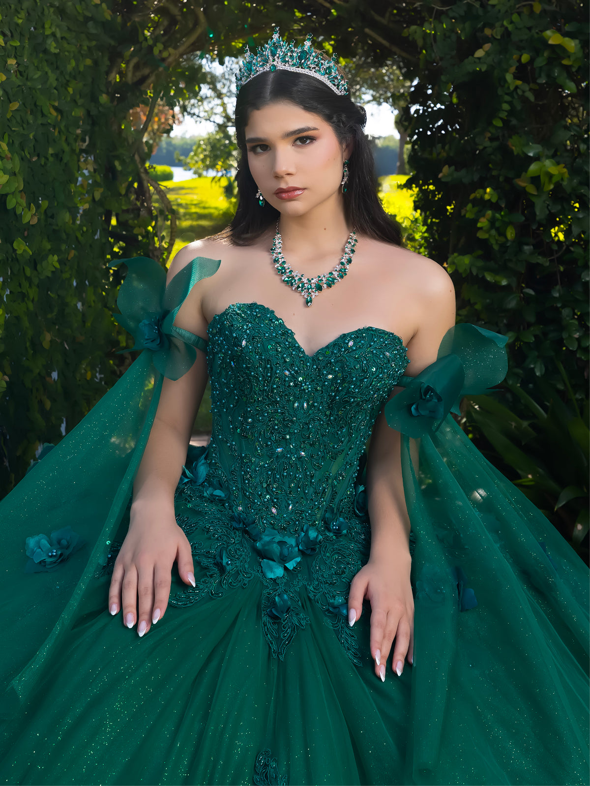 A woman wearing an emerald green strapless ball gown with sequined embellishments, floral appliqué, and sheer draped fabric, posed outdoors in front of a leafy green archway. / This ball gown features a floral-embellished bodice and a full tulle skirt with cascading 3D flowers, finished with a decorative bow at the back.