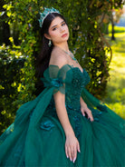 A young woman wearing a voluminous emerald green quinceañera ball gown with off-the-shoulder sheer fabric, beaded bodice, and matching green tiara and earrings, posed outdoors against a leafy green background. / This ball gown features a floral-embellished bodice and a full tulle skirt with cascading 3D flowers, finished with a decorative bow at the back.