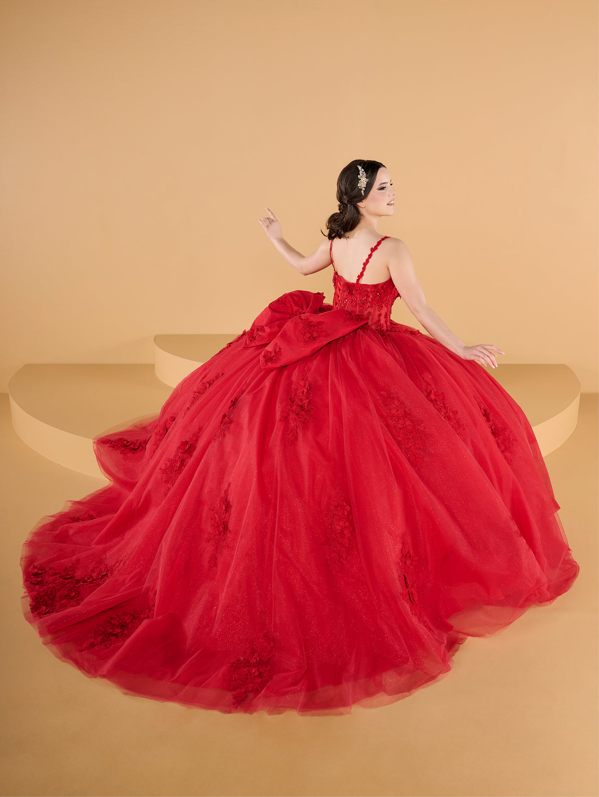 A woman wearing a voluminous red ball gown with floral lace details, posed on a beige studio set with curved platforms. / This ball gown features a fitted, embellished bodice with thin straps and a full skirt decorated with floral appliqué, finished with a bow at the back.