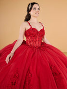 A young woman wearing a voluminous red ball gown with floral lace appliqué and a sheer embroidered bodice, posed against a warm beige studio background. / This ball gown features a fitted, embellished bodice with thin straps and a full skirt decorated with floral appliqué, finished with a bow at the back.